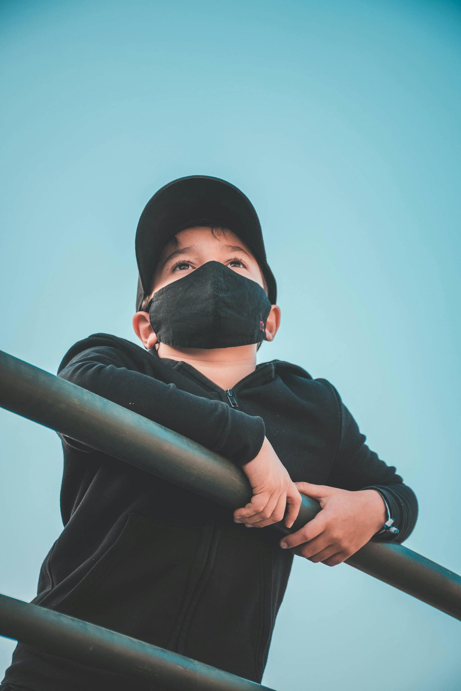 Low angle of male in black hoodie and cap with protective mask leaning on metal fence under blue cloudless sky in daylight and looking away